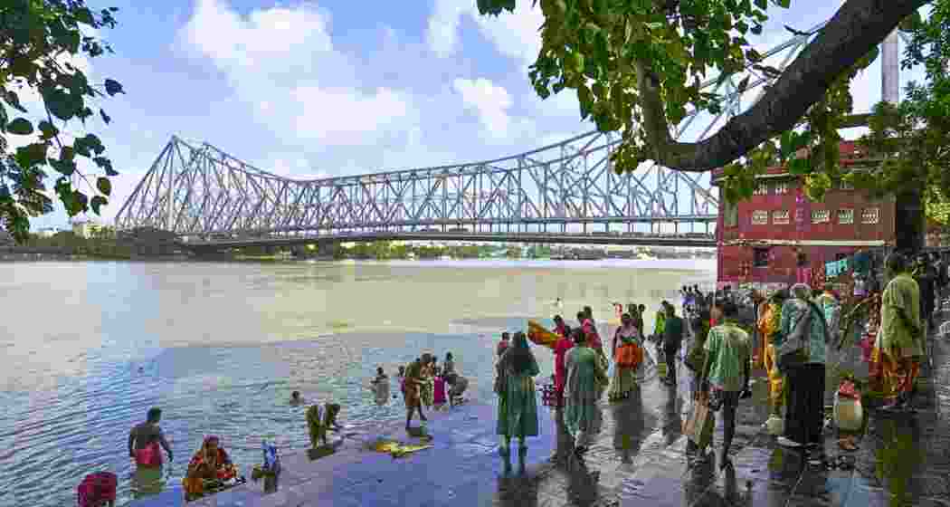 People gather along a Ganga ghat in Kolkata, West Bengal, with the Howrah Bridge visible in the background.