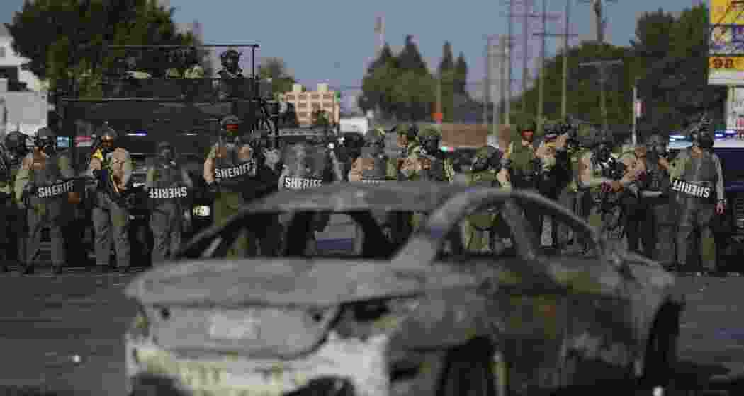 A National Guard team stands behind a charred vehicle in Los Angeles after immigration raid protests turned violent, with demonstrators clashing with federal agents and torching property on Saturday.